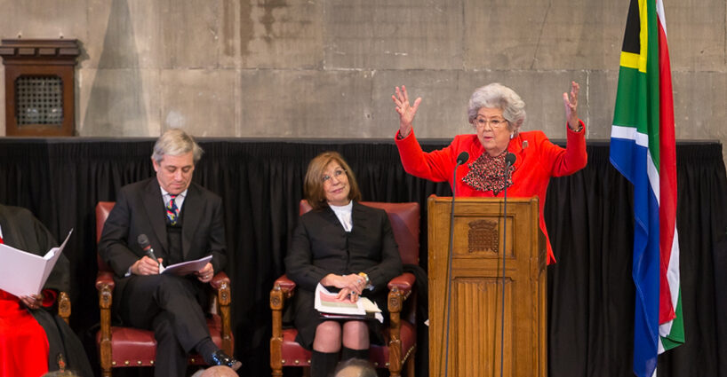 Baroness Betty Boothroyd wearing a red suit, standing behind a podium with arms raised.