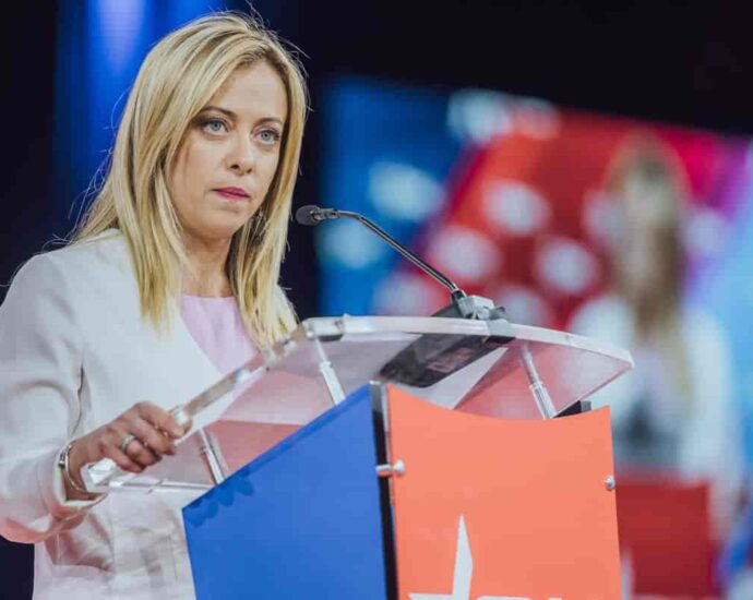 Image of the Prime Minister of Italy, Giorgia Meloni, standing behind a podium at a conference. The blonde haired woman is wearing a white long sleeved dress.