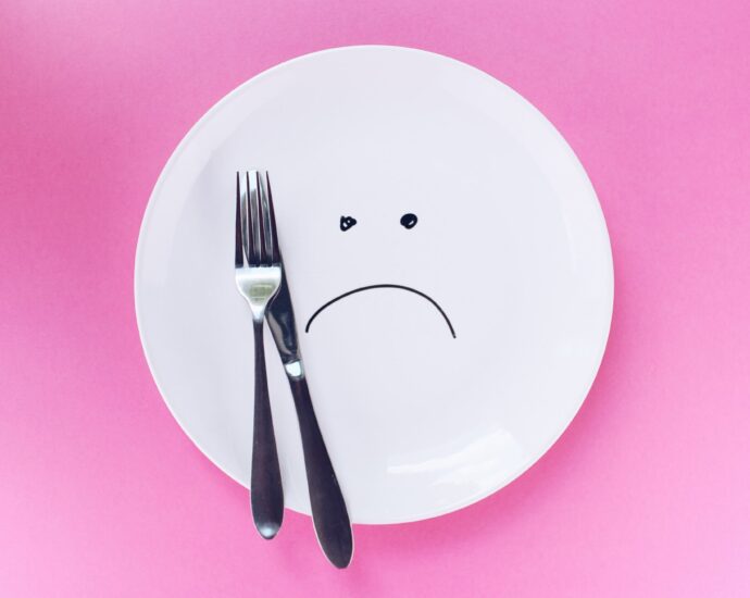 A plate with a sad face drawn on it, with a knife and fork, on a pink background.