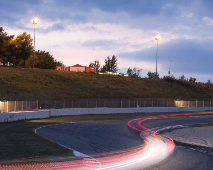 racing track at twilight, as the lights start to come on. a red blur is seen to show a car speeding past. formula one strategy is key to ensure high performance in all conditions.
