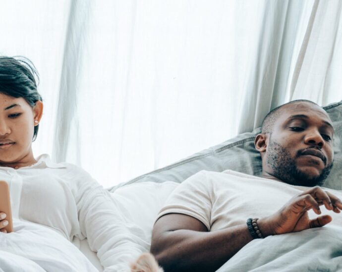 Image shows a woman and a man lying next to each other in bed on their mobile phones