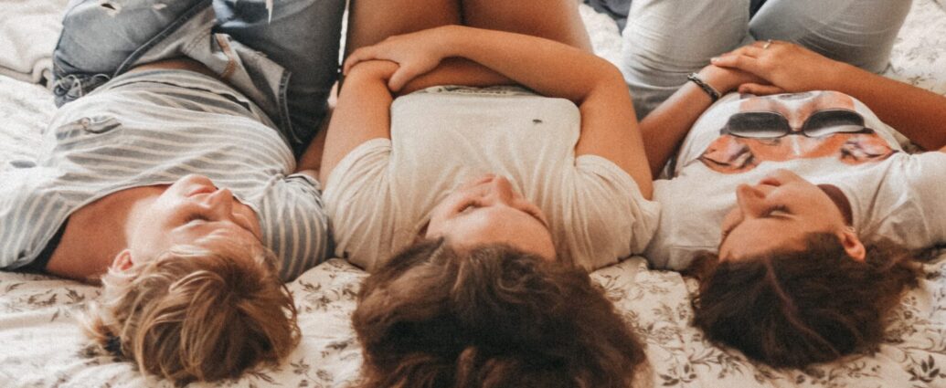 Image shows three teenage girls lying on a bed side-by-side.