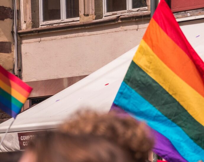 A rainbow pride flag is waving above a crowd of people. Flags like these can be seen in Heartstopper season two