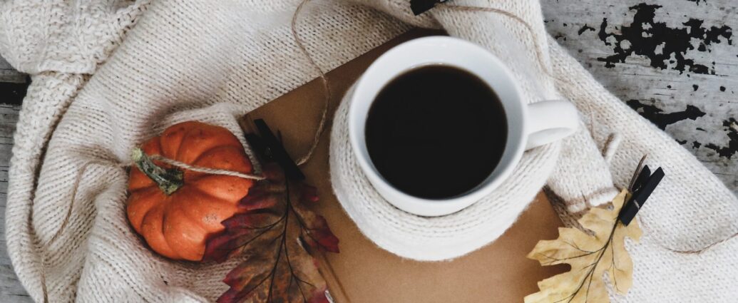 Autumn leaves, a white ceramic mug, a pumpkin, and a book sitting on a sweater.