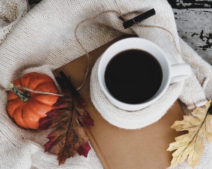 Autumn leaves, a white ceramic mug, a pumpkin, and a book sitting on a sweater.