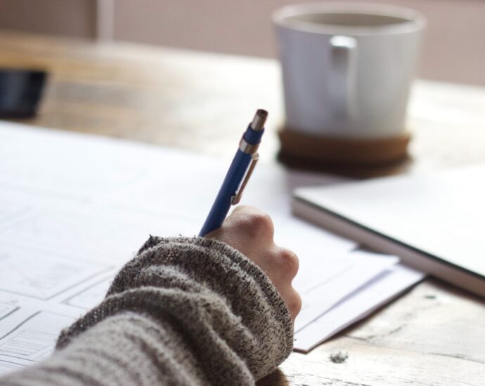 Person writing on brown wooden table near white ceramic mug. My journalism journey.