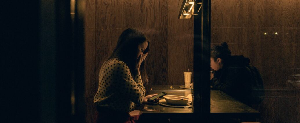 A girl sat at a restaurant table looks at her phone in shock.