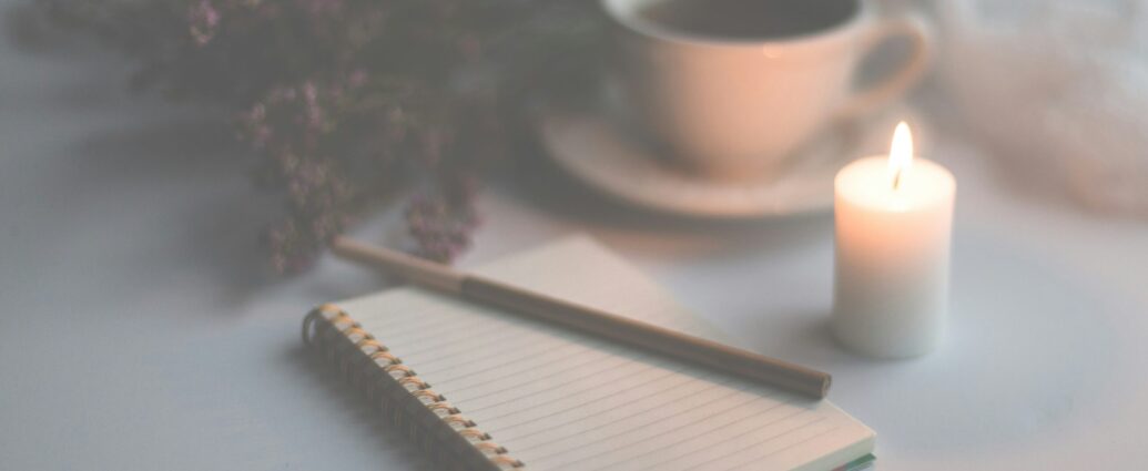 Image shows an open journal and pen, surrounded by a lit candle, cup of tea and dried flowers.