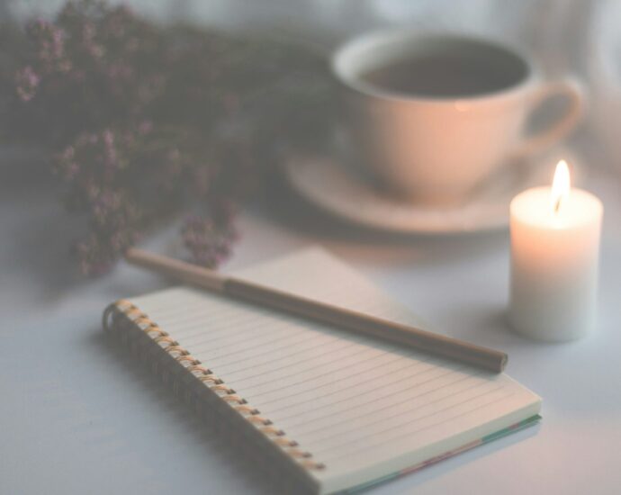 Image shows an open journal and pen, surrounded by a lit candle, cup of tea and dried flowers.