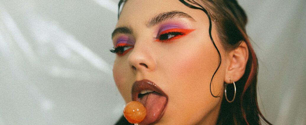A woman with Y2K style waved hair, brown lipstick and brightly coloured eye makeup licks a lollypop.