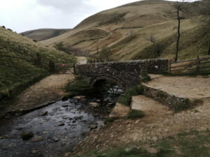 A green hilly landscape in the background, with a cobbled bridge over a river.