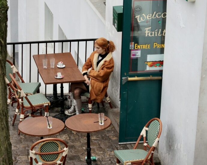 A woman with ginger hair and brown coat sitting at a table outside a restaurant.