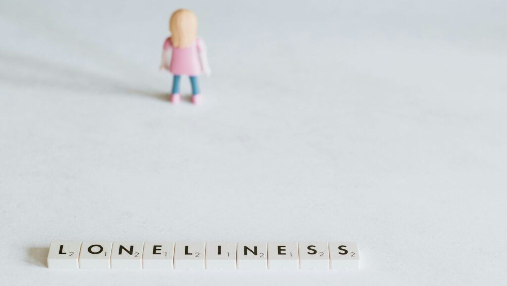 A girl figurine in pink top and blue trousers next to scrabble letters spelling loneliness.