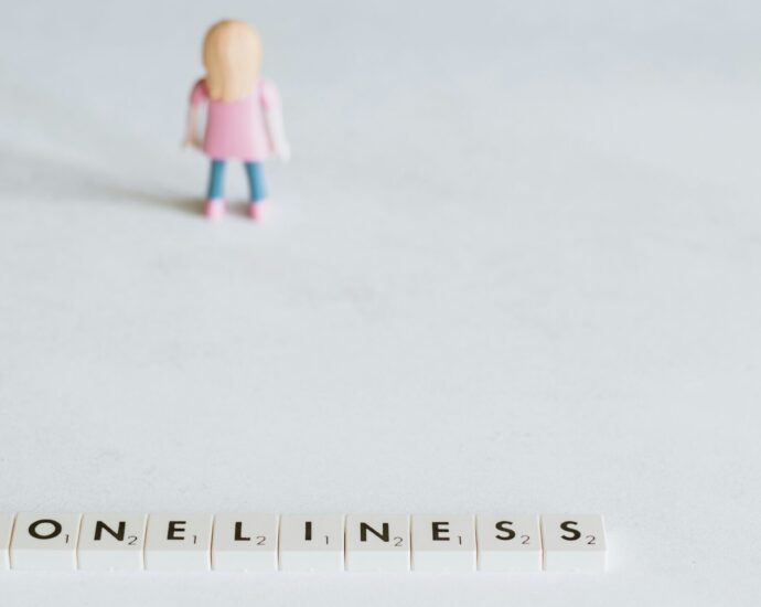 A girl figurine in pink top and blue trousers next to scrabble letters spelling loneliness.
