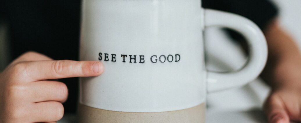 A finger is seen pointing at the words "see the good" engraved on a white mug.