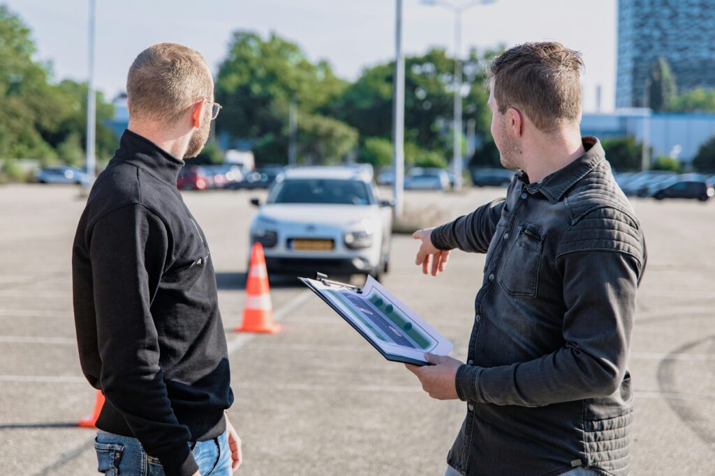 Driving instructor in a car park pointing to to a traffic cone with student and car in the background.