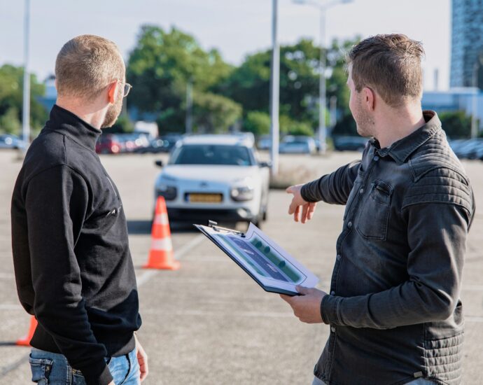 Driving instructor in a car park pointing to to a traffic cone with student and car in the background.