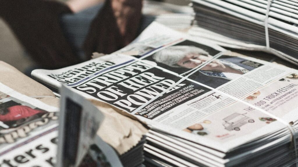 A stack of newspapers to represent the theme of 'journalism.' The image is blurred but the front page reads 'Stripped of her power' and shows Teresa May.
