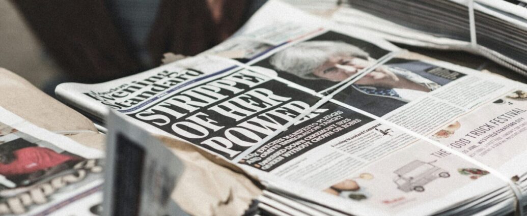 A stack of newspapers to represent the theme of 'journalism.' The image is blurred but the front page reads 'Stripped of her power' and shows Teresa May.