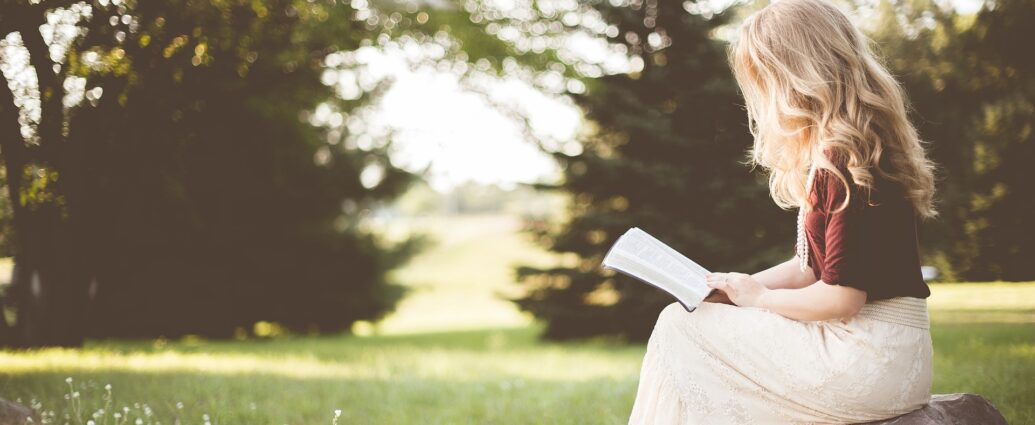 Girl sitting reading a book outside with grass and trees in the background potentially sunrise on the reaping