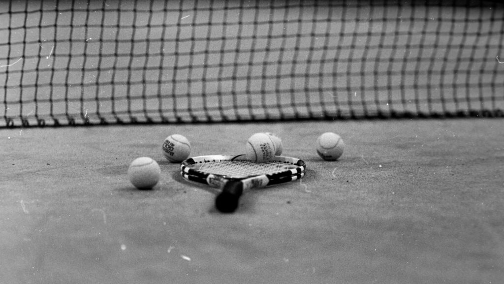 Black and white picture of a tennis racket lying on the ground surrounded by tennis balls.