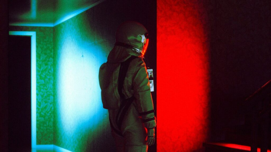 Person wearing a space suit standing in a dark room with the colors red and green reflecting on the walls.