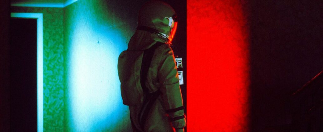 Person wearing a space suit standing in a dark room with the colors red and green reflecting on the walls.